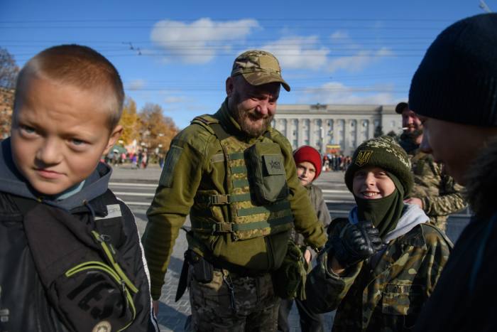 Young boys stand in the sunshine, next to a soldier who is smiling