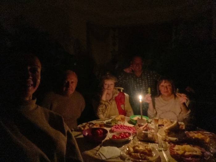 People are gathered around a dinner table. The room is dark, the only light from a single candle